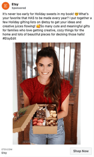 A smiling woman in a red top holds a box of assorted holiday treats and gifts, standing indoors in front of a white curtain.