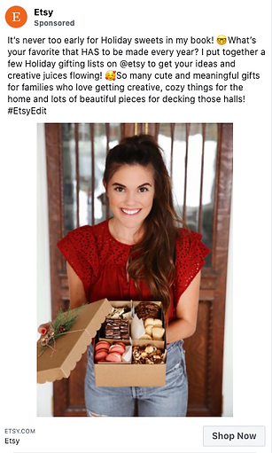 A smiling woman in a red top holds a box of assorted holiday treats and gifts, standing indoors in front of a white curtain.