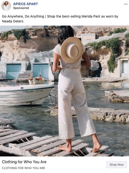 A woman stands on a wooden dock by the water, holding a hat behind her back and facing away, wearing light-colored pants and a top. Small boats and buildings are visible in the background.