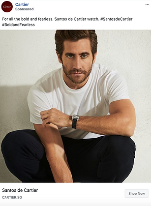 A man in a white t-shirt and dark pants sits against a light wall, wearing a Santos de Cartier watch on his left wrist.