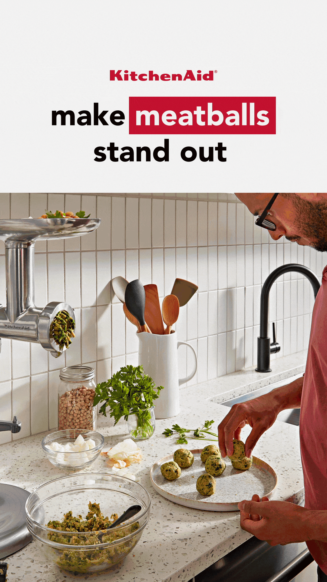 A person shapes meatballs by hand in a modern kitchen, with a meat grinder, a bowl of mixture, herbs, and kitchen utensils visible on the counter.