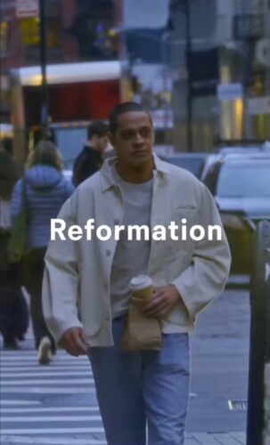 A person in a light jacket holding a coffee and paper bag walks across a city street. The word "Reformation" is overlaid on the image.