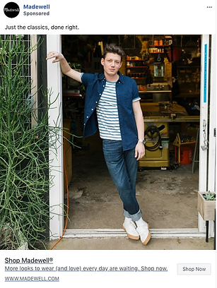 Person wearing a blue short-sleeve shirt over a striped t-shirt, jeans, and white shoes stands in a garage doorway with one arm resting on a tall plant.