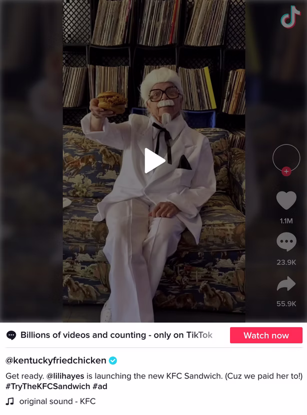 A person dressed as Colonel Sanders sits on a patterned sofa, holding up a sandwich, in a promotional TikTok video for KFC.
