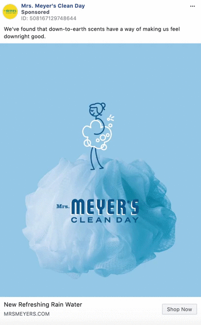 A blue bath pouf with the Mrs. Meyer's Clean Day logo and a simple line drawing of a person washing with soap bubbles. Text above reads: "New Refreshing Rain Water.