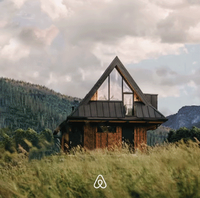 A-frame cabin with large windows stands in a grassy field, surrounded by trees and mountains under a cloudy sky. Airbnb logo is visible at the bottom.