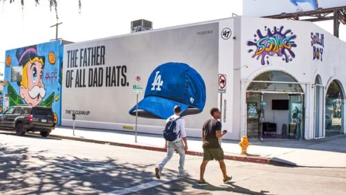 Two people walk past a building with colorful murals and a large billboard advertising a blue LA baseball cap that reads, “The father of all dad hats.”.
