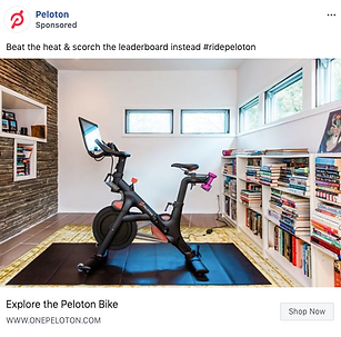 A Peloton exercise bike is positioned on a mat in a modern, well-lit room with bookshelves lining the walls and large windows above.