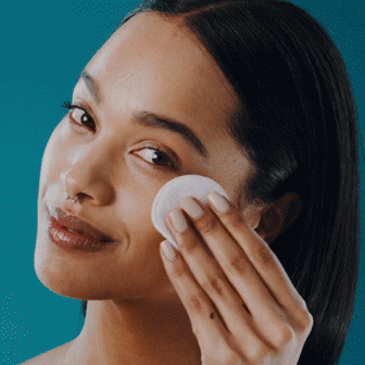 A woman with straight dark hair uses a cotton pad to apply or remove skincare product on her cheek against a teal background.