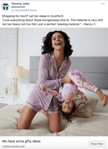 A woman in lavender pajamas sits on a bed smiling, holding a laughing child upside down in playful pajamas.