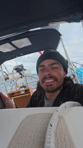 A person wearing a beanie and jacket smiles while sitting at the helm of a sailboat on open water under a cloudy sky.