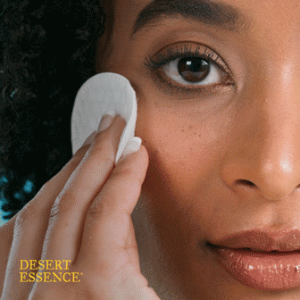A person uses a cotton pad to cleanse their cheek. The words "Desert Essence" appear in the lower left corner.