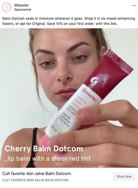 A woman holds a tube of Glossier Cherry Balm Dotcom lip balm close to the camera, highlighting the product label.