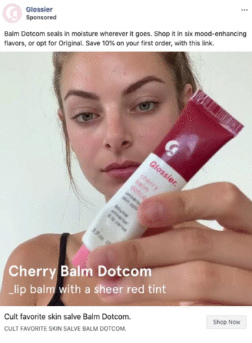 A woman holds a tube of Glossier Cherry Balm Dotcom lip balm close to the camera, displaying the product label.