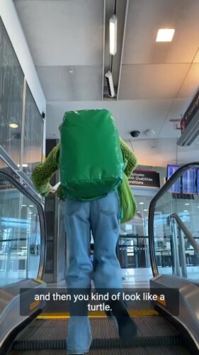 Person wearing a large green backpack and blue jeans rides an escalator at an airport; digital sign and flight boards visible in the background.