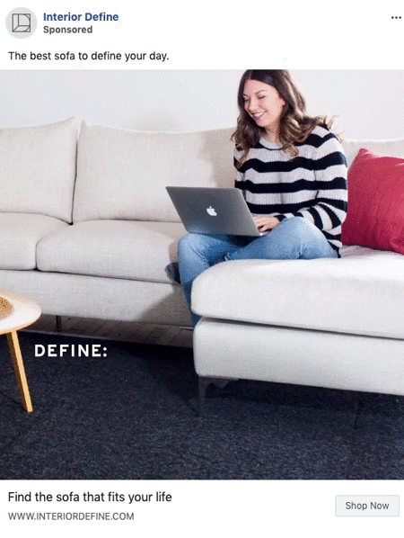 A woman sits on a light-colored sofa, smiling and using a laptop, with a red cushion beside her.