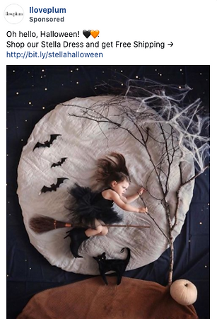 A young girl in a black dress poses as a witch on a broomstick, surrounded by bats, a black cat, and Halloween-themed decorations on a moon-like backdrop.