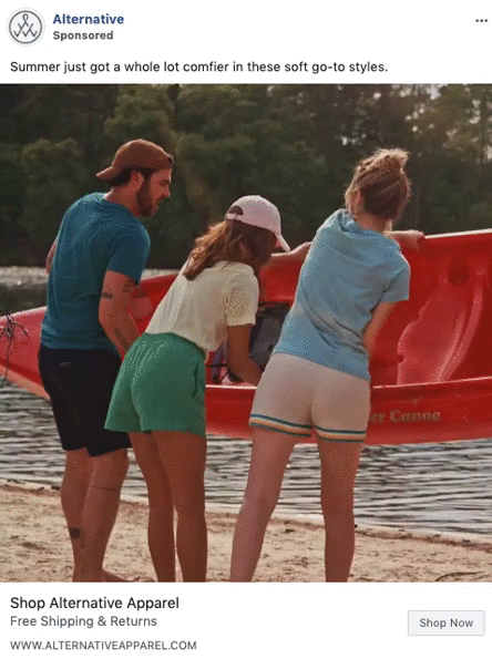 Three people on a sandy shore lift a red kayak together. All are dressed in casual summer clothing. Trees and water are visible in the background.