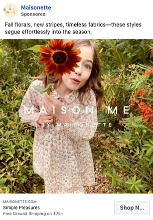 Young girl in a floral dress holds a large orange flower towards the camera while standing outdoors among green plants.