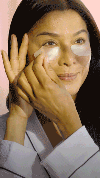 A person applies under-eye patches while looking in the mirror, wearing a light-colored robe.
