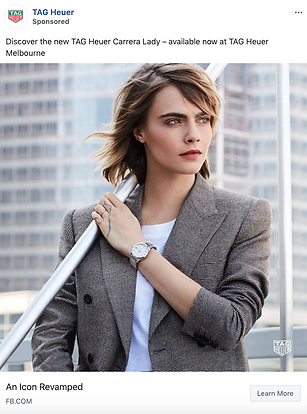 A woman in a gray blazer and white shirt stands outdoors by a railing, wearing a wristwatch. Urban buildings are visible in the background.