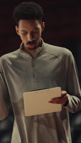 A man in a light gray pullover holds a clipboard and looks down, appearing focused, with a dark blurred background.