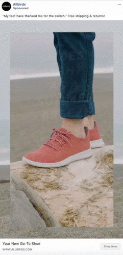 A person standing on a driftwood log at the beach wearing coral-colored Allbirds sneakers and rolled-up jeans.