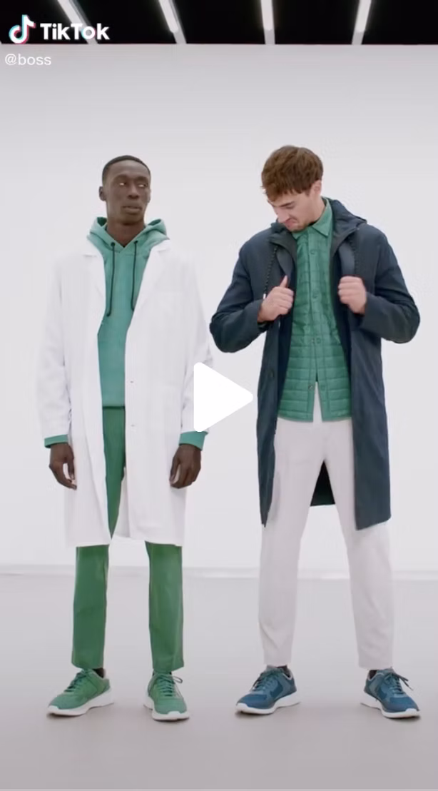 Two men stand side by side in a studio, both wearing layered green and white outfits with sneakers, looking at each other against a plain white background.