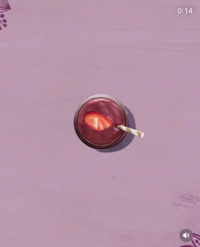 A glass of purple smoothie with a paper straw and a strawberry slice on top sits on a light purple surface.