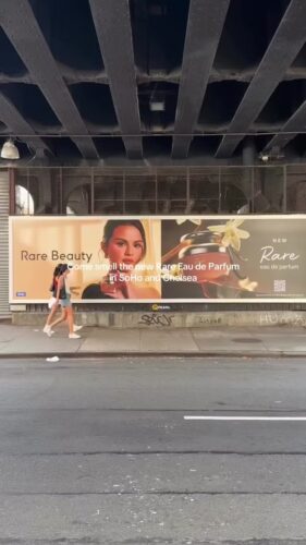 A person walks past a billboard under a bridge promoting Rare Beauty's new Rare Eau de Parfum in SoHo and Chelsea, featuring a close-up of a woman's face and a perfume bottle.