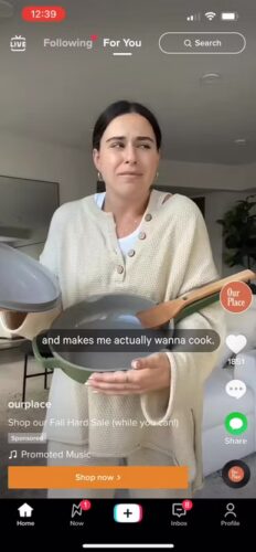 A woman in a cream sweater holds a gray pan with a lid and spatula. Text on-screen reads, "and makes me actually wanna cook." App interface elements are visible.
