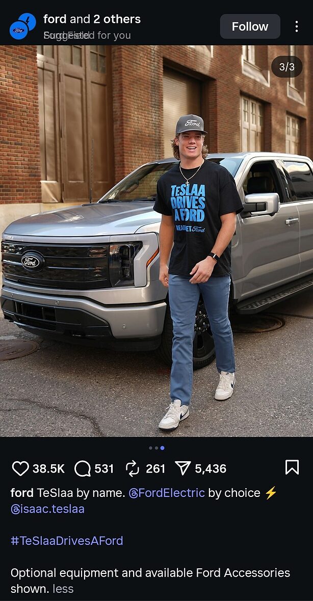 A man wearing a "TeSlaa Drives A Ford" T-shirt and cap smiles while standing in front of a silver Ford pickup truck on a city street.
