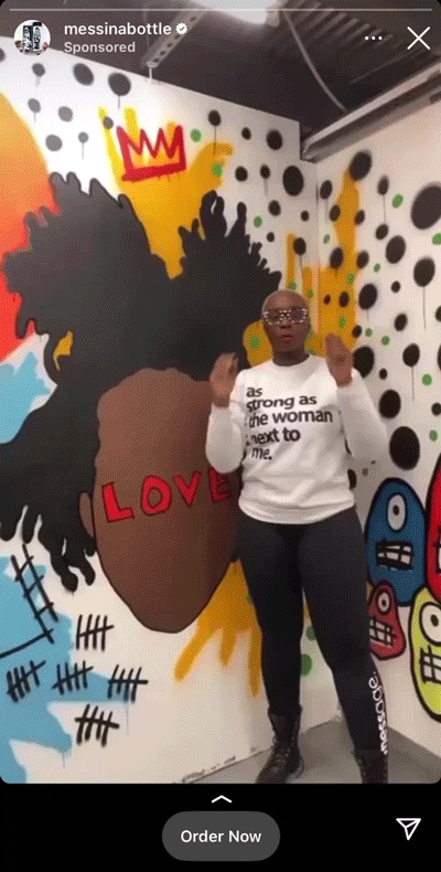A person stands in front of a colorful mural with abstract faces, wearing glasses and a white sweatshirt that reads, "As strong as the woman next to me.