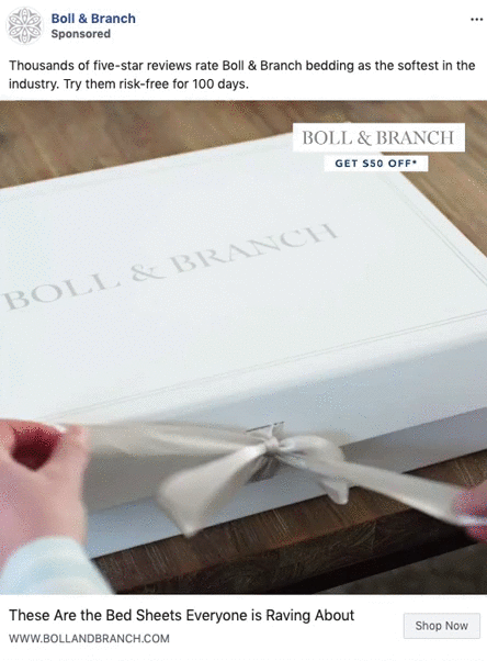 A hand unties a ribbon on a white Boll & Branch box placed on a wooden surface. The box displays the brand's name in gray text.
