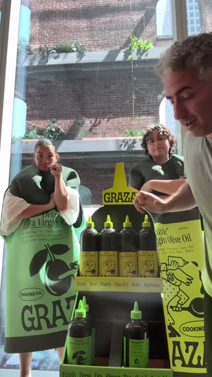 Two people wear costumes resembling bottles of olive oil, while a third person stands beside a display of large olive oil bottles indoors.