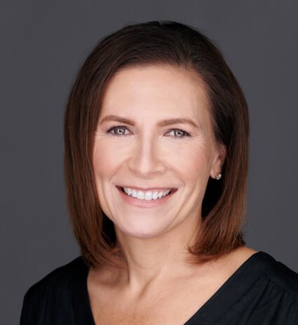Woman with straight, shoulder-length brown hair, smiling, wearing a black top, against a plain gray background.