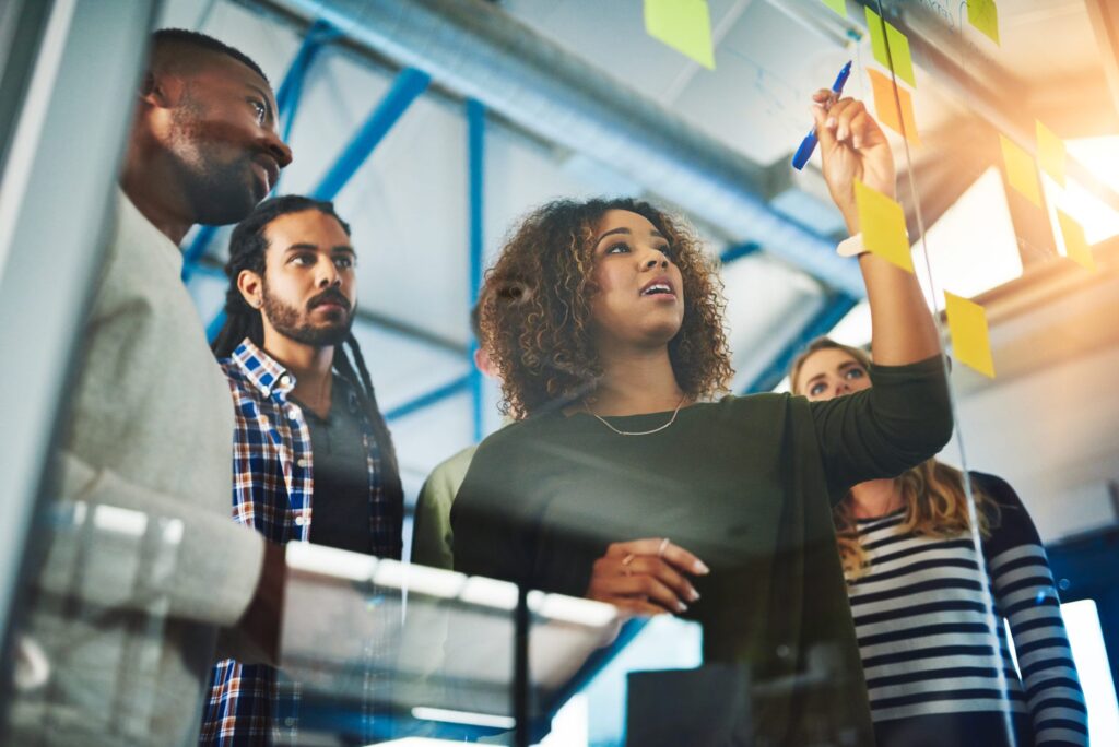 A group of people stands around a glass board covered with sticky notes as a woman writes or draws on it with a blue marker.