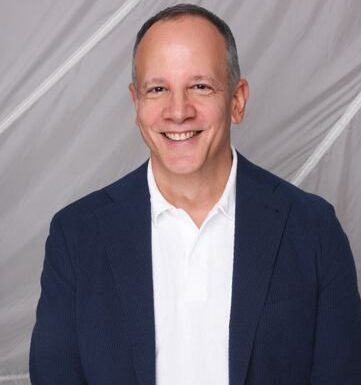 A man in a navy blazer and white polo shirt smiles at the camera against a light-colored, draped background.