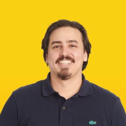 A man with dark hair, a mustache, and a beard smiles at the camera while wearing a navy polo shirt against a solid yellow background.