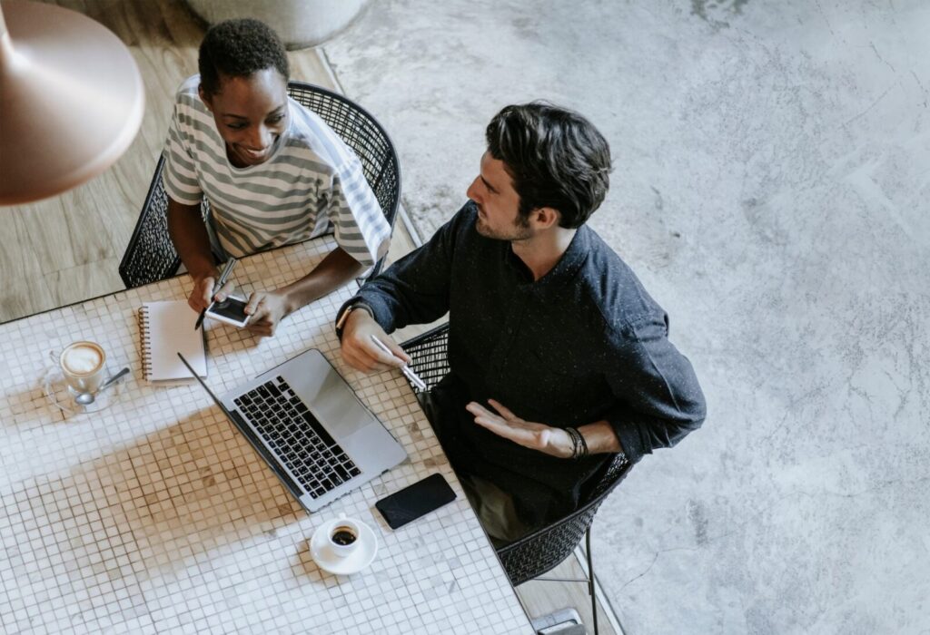 Two people sit at a table with a laptop, notebooks, and coffee cups, engaged in conversation and collaborating on AI search marketing strategies.