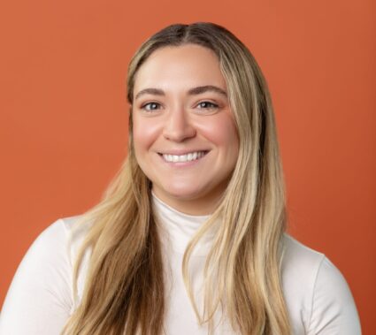 Woman with long blonde hair wearing a white turtleneck, smiling in front of an orange background.