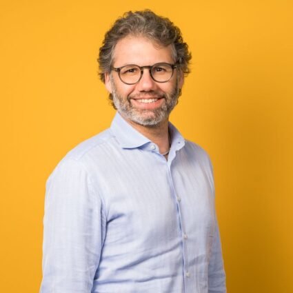 Man with curly hair and beard wearing glasses and a light blue shirt, standing and smiling in front of a solid yellow background.