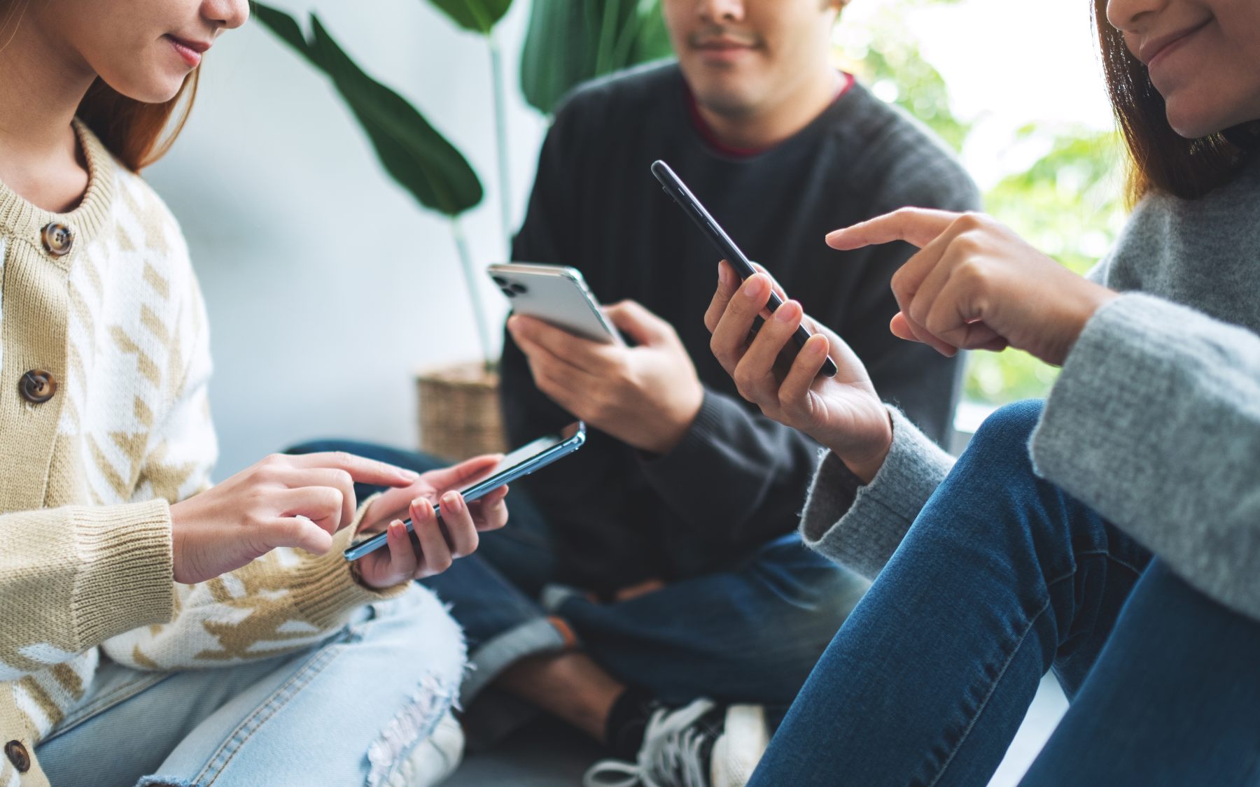 Three people sitting together on the floor, each using a smartphone, with plants visible in the background.
