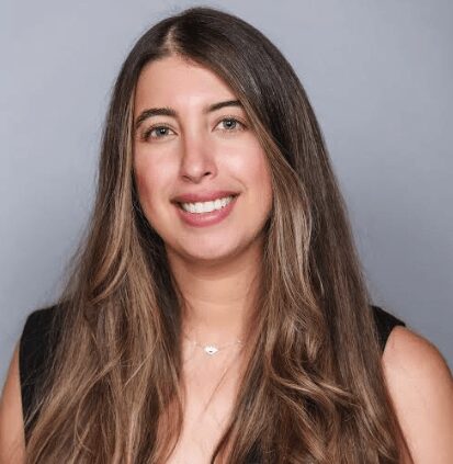 Woman with long brown hair wearing a black top and a necklace, smiling at the camera against a plain gray background.