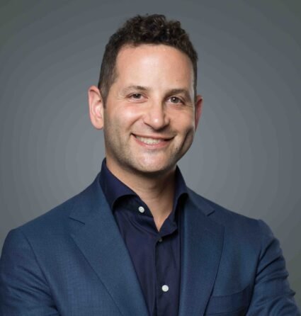 A man in a blue suit and dark shirt smiles at the camera against a plain gray background.