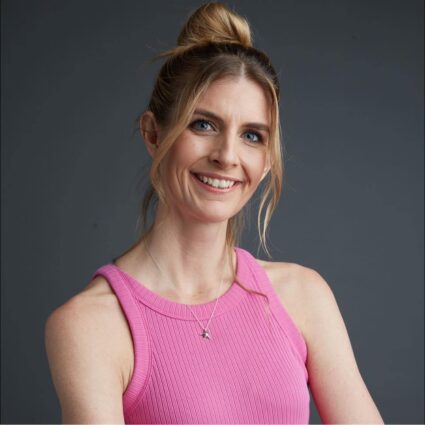 Woman with blonde hair in a bun, wearing a sleeveless pink top and a necklace, smiling against a plain gray background.