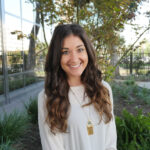 A woman with long, wavy brown hair and a white blouse smiles while standing outside near greenery and a building with reflective windows.