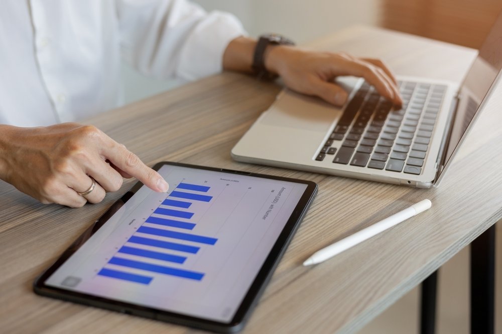 Marketing professional reviewing performance metrics on a tablet bar chart alongside a laptop.