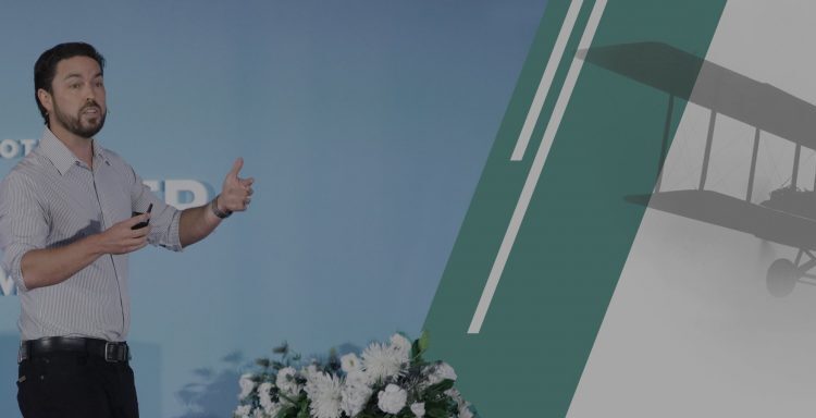 A man stands on stage giving a presentation at the Wpromote Challenger Summit, with a floral arrangement and a vintage airplane graphic in the background.