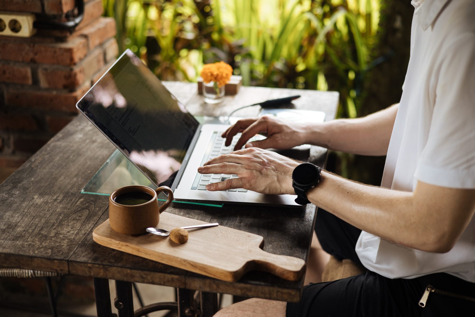 Person typing on a laptop at an outdoor table with a cup of coffee, a spoon, and a wooden cutting board in front of them; greenery and a brick wall in the background.